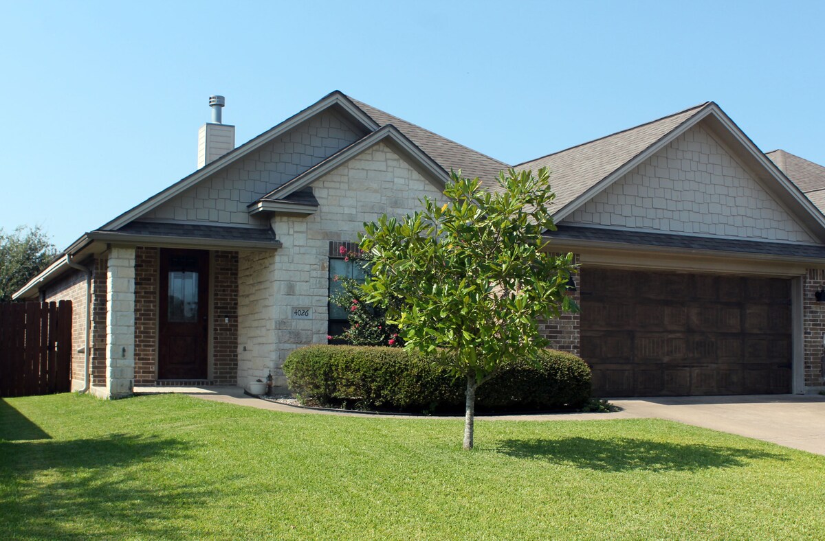 The charming exterior of the home showcases a blend of brick and stone materials, complemented by a well-manicured lawn. A symmetrical arrangement of windows is visible, along with a two-car garage and a welcoming entrance framed by landscaping.