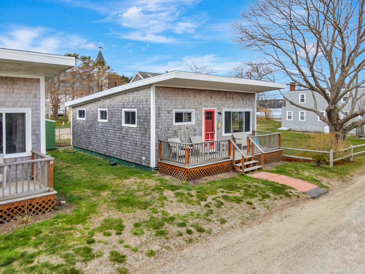 A charming cottage exterior is featured, showcasing a light-colored shingle facade and a vibrant pink door. A wooden porch is visible, offering steps that lead to the grassy front yard. Nearby trees and other structures contribute to the serene surroundings.