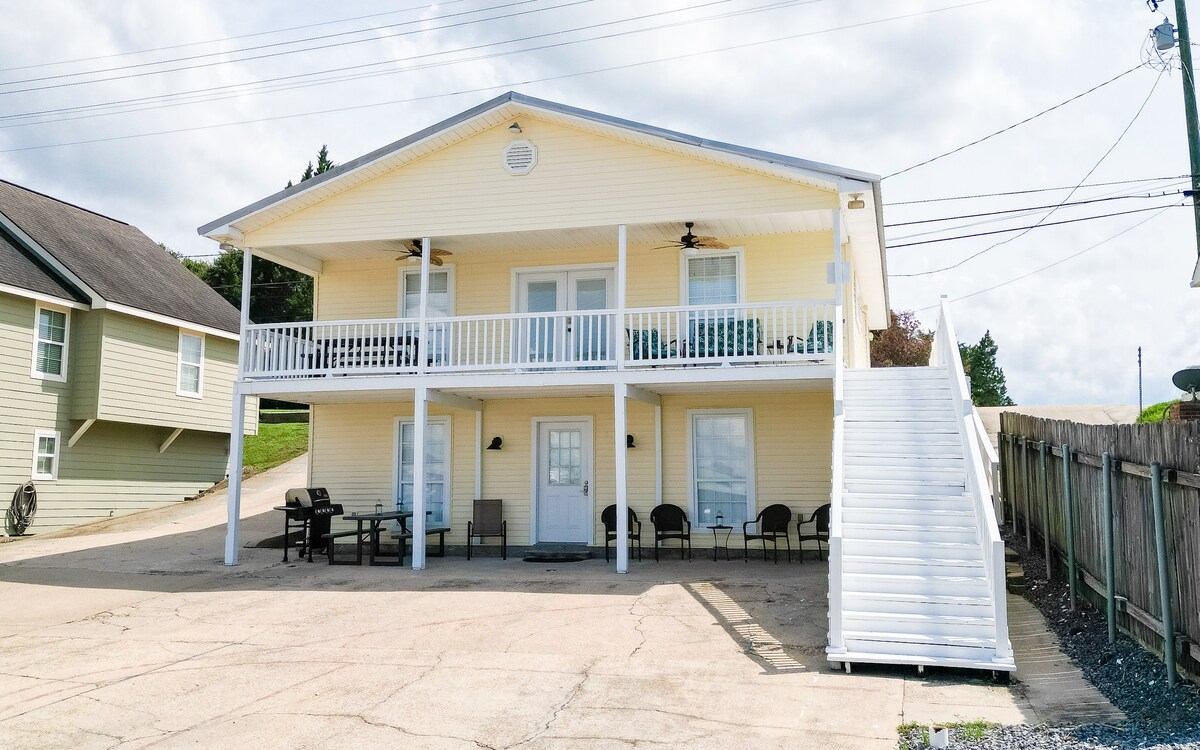 A two-story yellow rental unit features a broad front porch with several chairs and tables. The entrance is flanked by glass doors, while a staircase leads to the upper level. The surrounding area includes a paved driveway and greenery in the background.