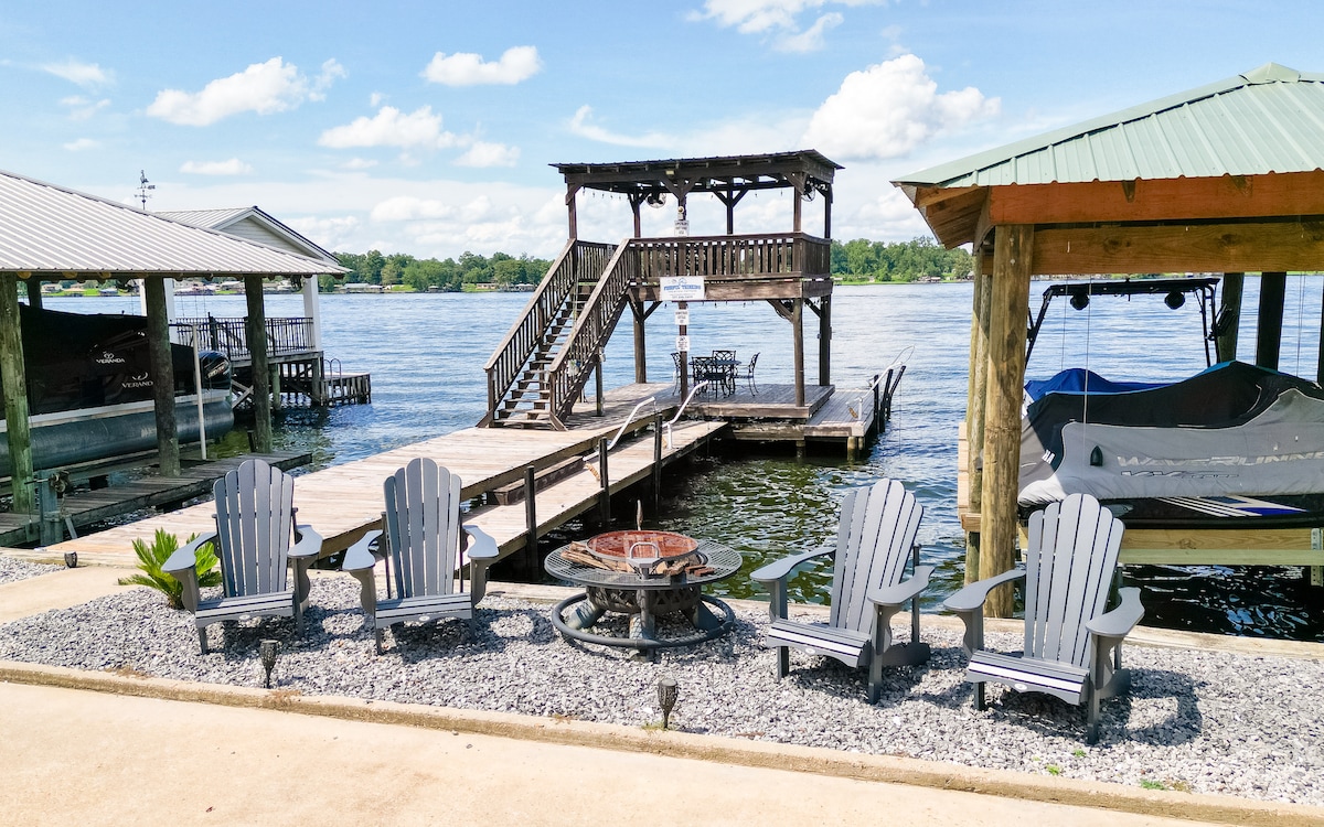 An outdoor area features a private pier with a multi-level structure overlooking the water. Four Adirondack chairs are arranged around a fire pit on a pebbled surface. A covered boat slip with a canopy is visible nearby, surrounded by calm waters and a clear sky.