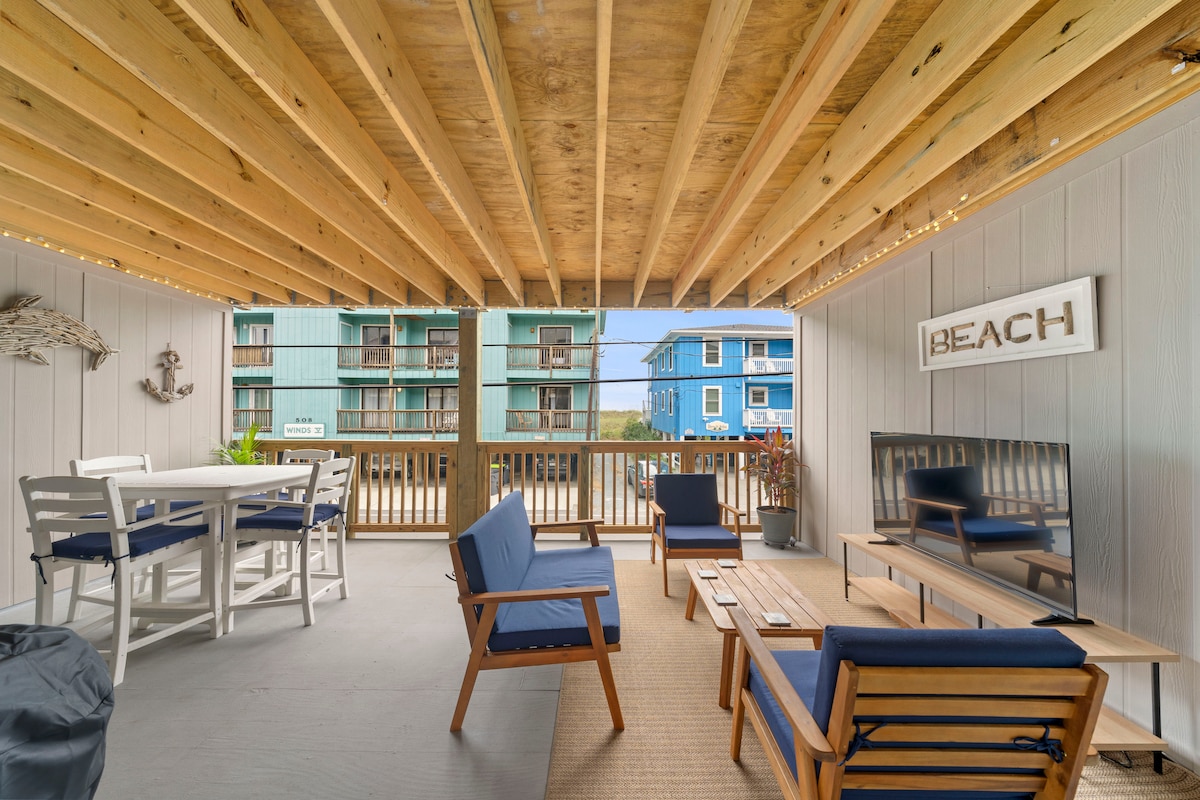 An outdoor covered porch features wooden beams and ambient twinkle lights. A dining table with four chairs occupies one corner, while a comfortable seating area includes two blue armchairs and a low table. A wall-mounted TV is positioned near a 'BEACH' sign, with views of neighboring buildings.
