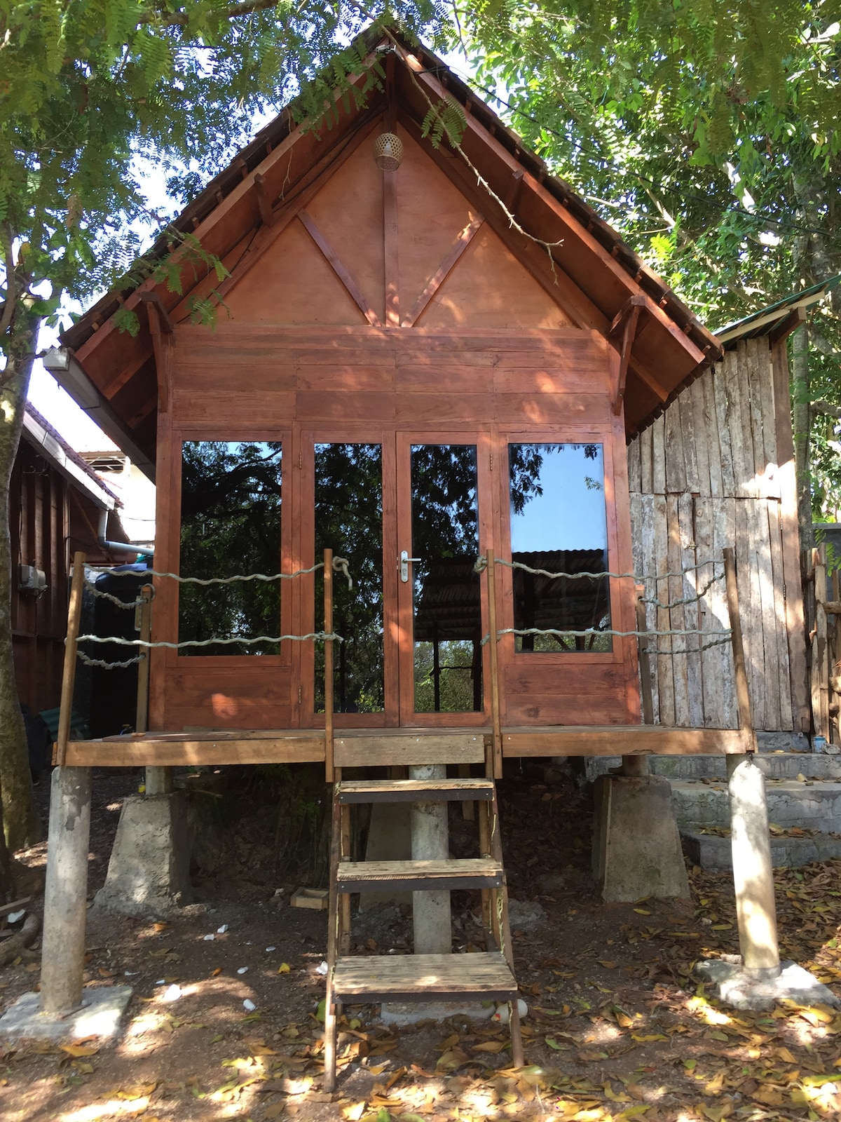 A wooden cabin with a distinctive A-frame roof is set on stilts, accessed by a wooden stairway. Large glass windows reflect the surroundings, enhancing the natural feel. The structure is surrounded by greenery, creating a serene environment.