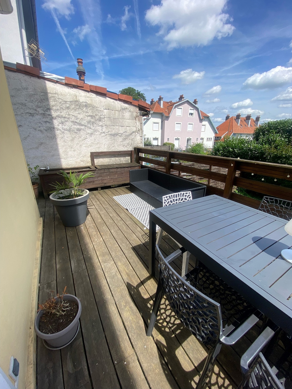 An outdoor terrace is presented, featuring a wooden deck and a rectangular dining table surrounded by chairs. Potted plants are visible, adding a touch of greenery. The scene is brightened by blue skies scattered with clouds, complemented by nearby residential rooftops.