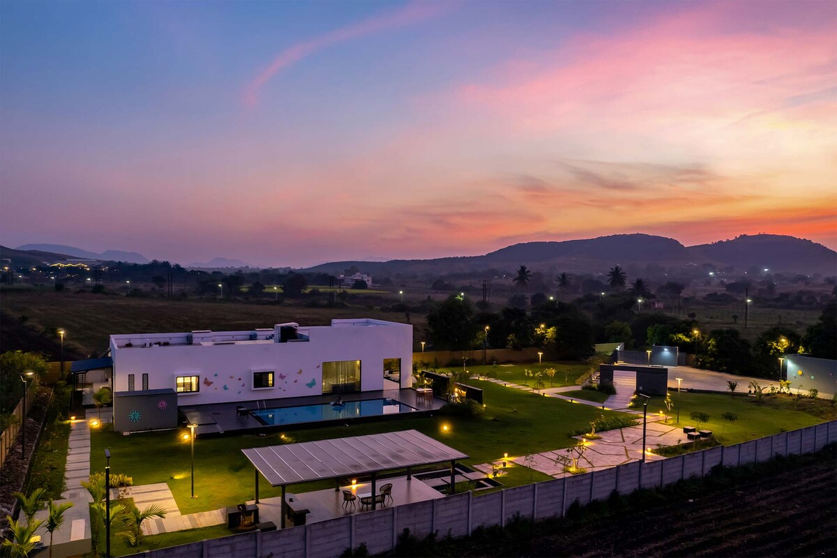 An aerial view captures the contemporary villa amid lush greenery, highlighting expansive outdoor spaces and a private lawn. The sky glows with shades of pink and orange at sunset, while the property is thoughtfully illuminated, emphasizing the inviting ambiance and panoramic surroundings.