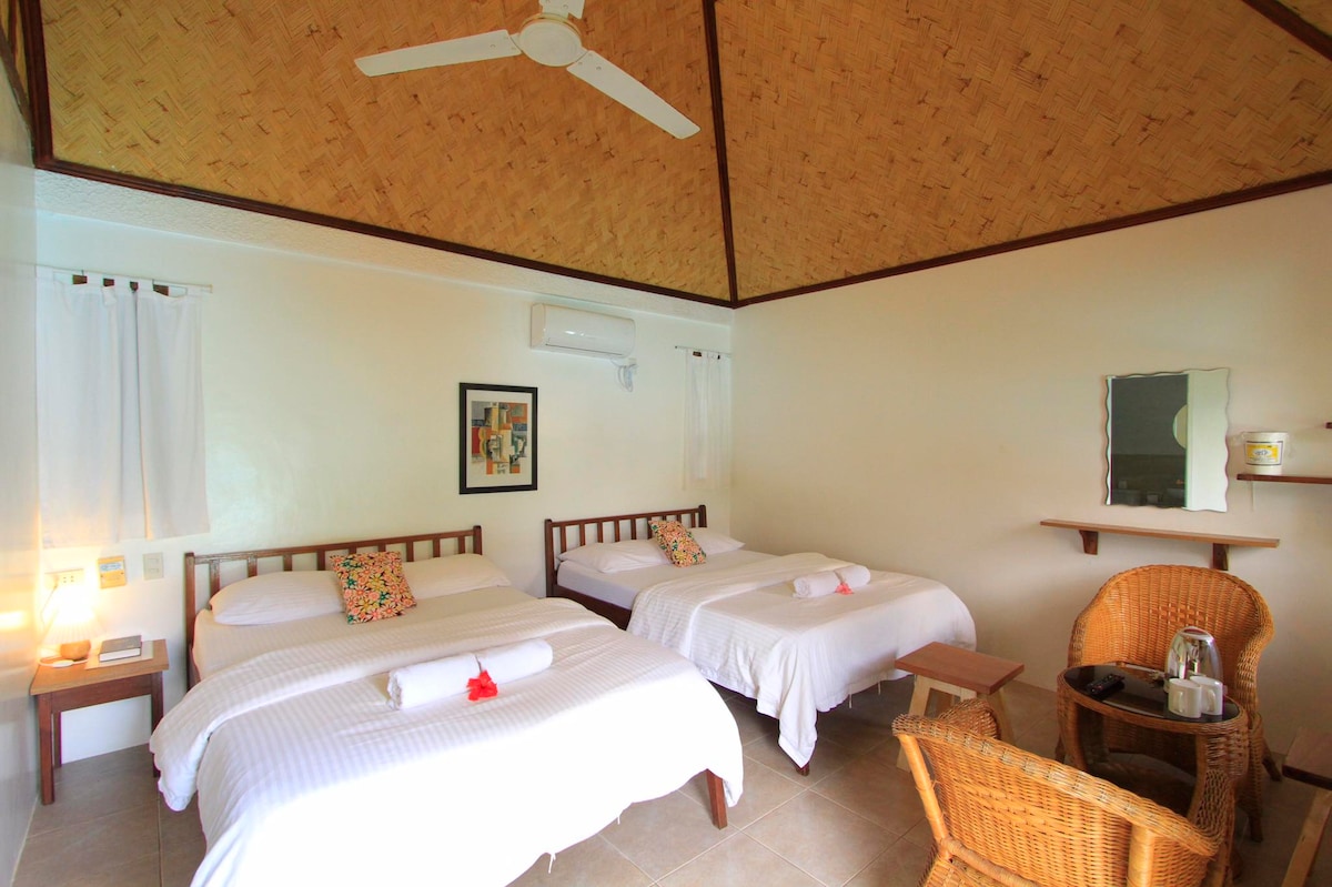 A bright sleeping area features two spacious beds adorned with rolled towels and floral accents. A chair made of natural fibers sits beside a wooden table, while a large mirror reflects light from nearby windows dressed in sheer curtains.