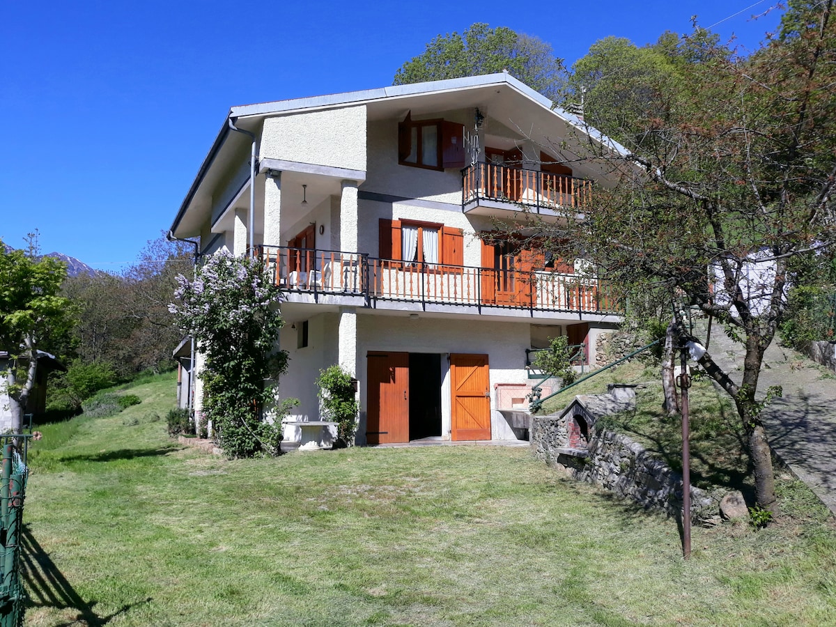 A spacious two-story house is surrounded by lush greenery. The exterior features light-colored walls and multiple wooden balconies with orange shutters. A well-kept lawn adds to the inviting setting, with ample outdoor space visible.