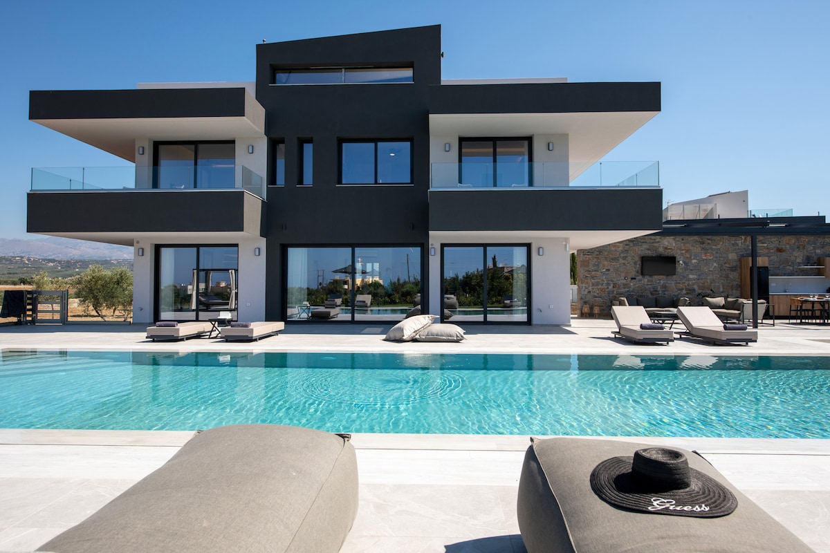 The villa's modern architecture is highlighted, showcasing multiple expansive glass doors and balconies on the second floor. A large private swimming pool reflects the clear blue sky, framed by elegant lounge chairs. The landscaped outdoor area provides a serene and inviting setting.