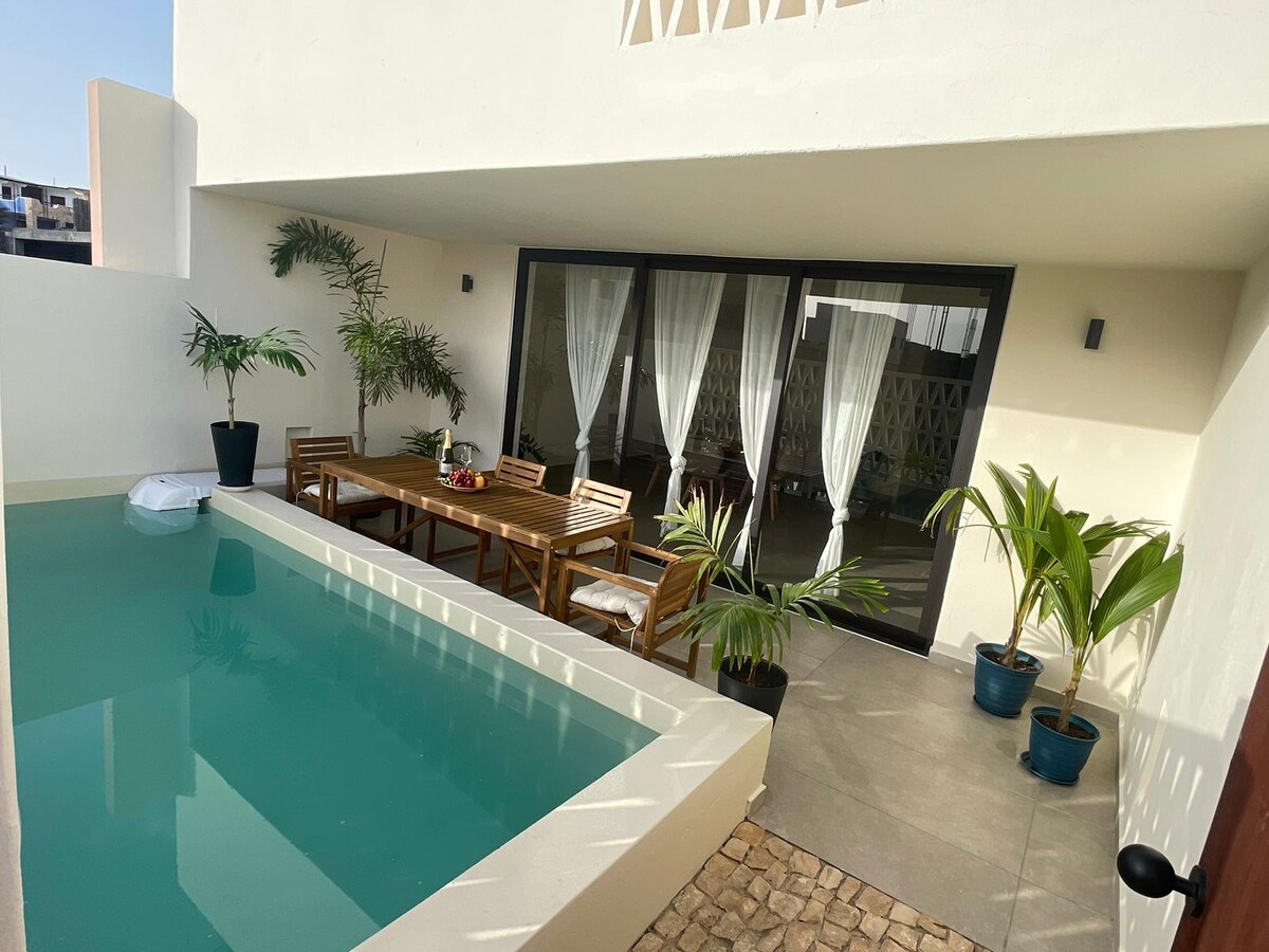 A private outdoor area features a turquoise pool beside a wooden dining table surrounded by several chairs. Potted plants add greenery, while large glass doors lead into the interior space, offering a connection between the indoor and outdoor environments.
