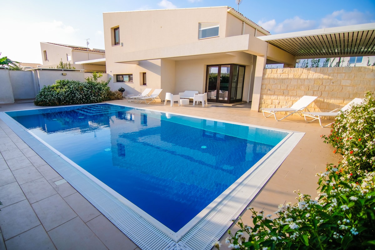 A modern villa features a private swimming pool surrounded by a spacious patio. Four lounge chairs are positioned beside the pool, and a neutral stone wall offers privacy. Lush greenery and flowering plants contribute to the inviting outdoor setting.