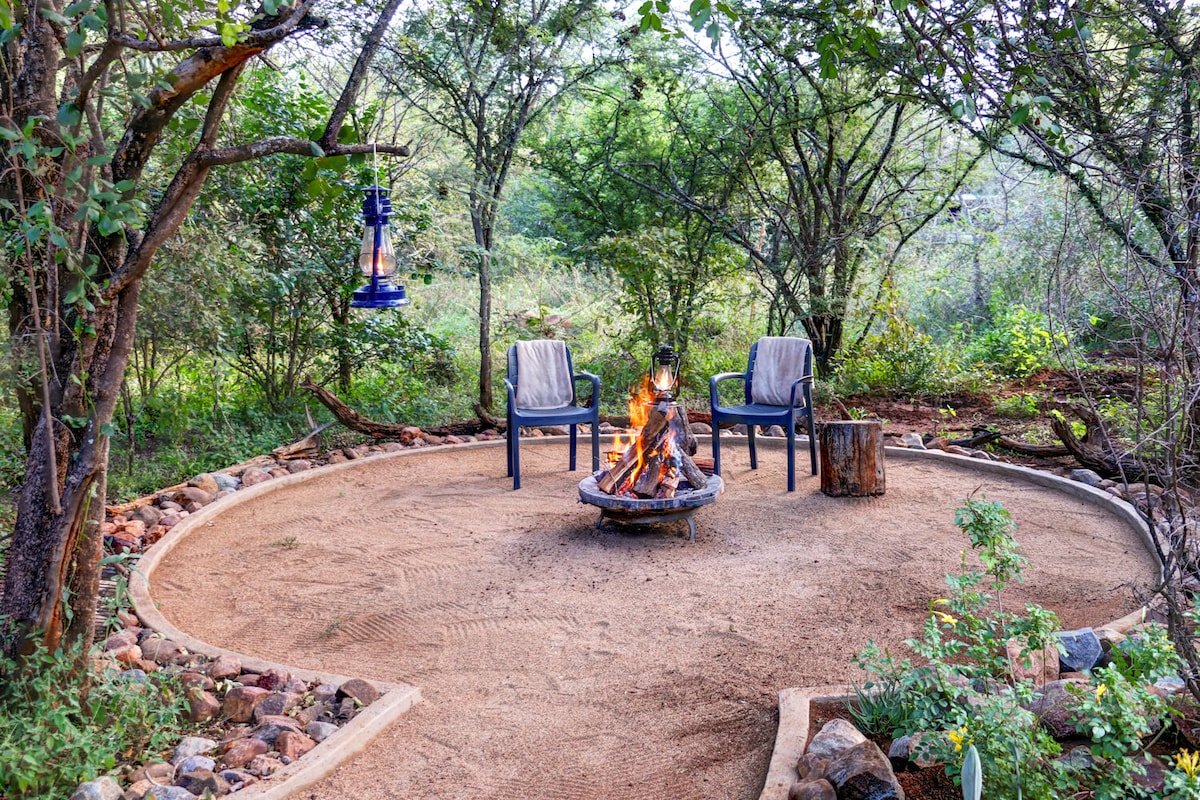 A cozy fire pit area is surrounded by a circular sandy space, featuring two chairs with soft blankets draped over them. Natural greenery and trees frame the setting, providing a serene environment for outdoor gatherings.