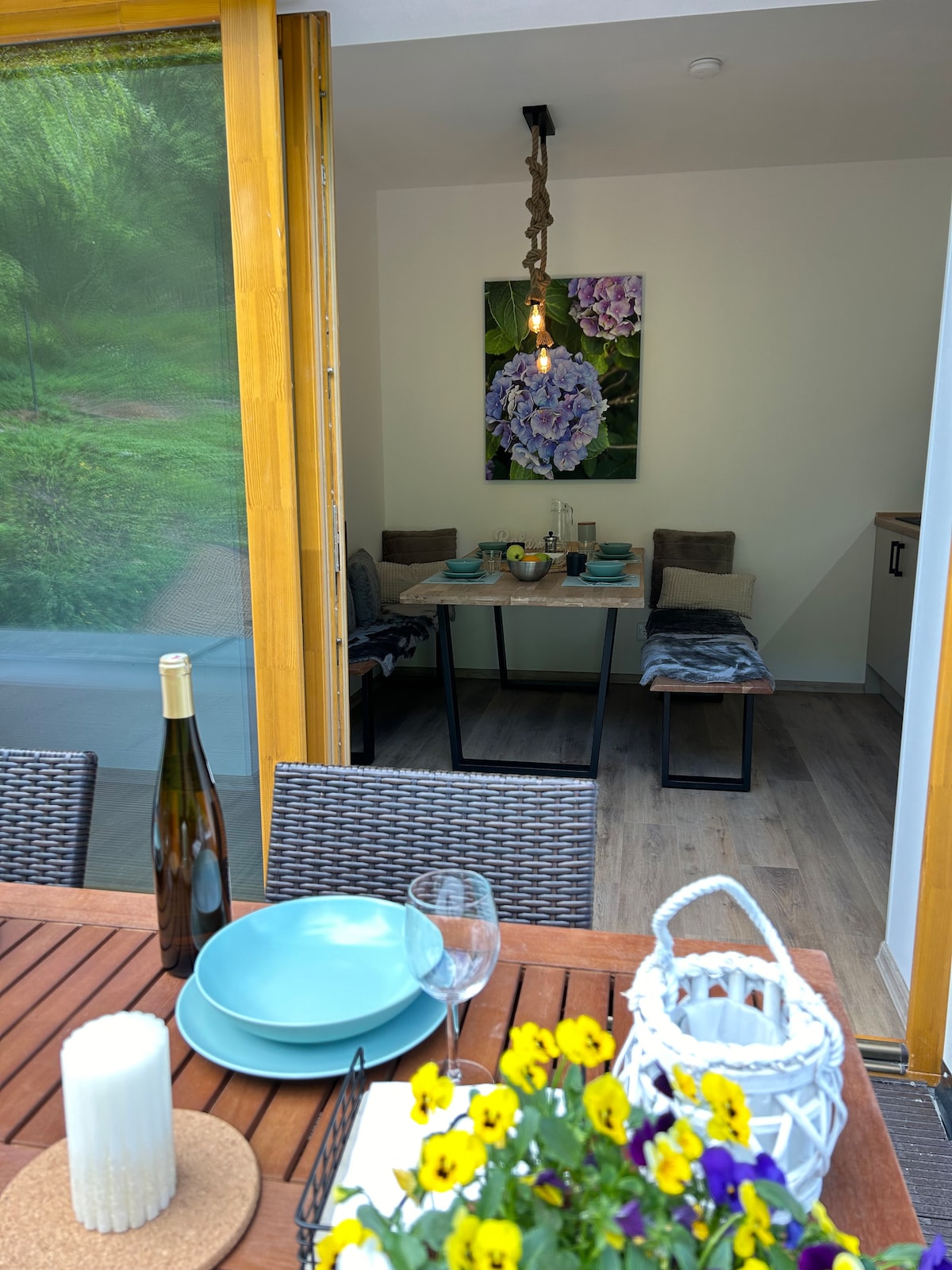 A dining table is set on a wooden outdoor terrace, featuring plates, drinks, and a floral centerpiece. In the background, a cozy dining area is visible through large glass doors, with a piece of wall art and a soft seating arrangement.