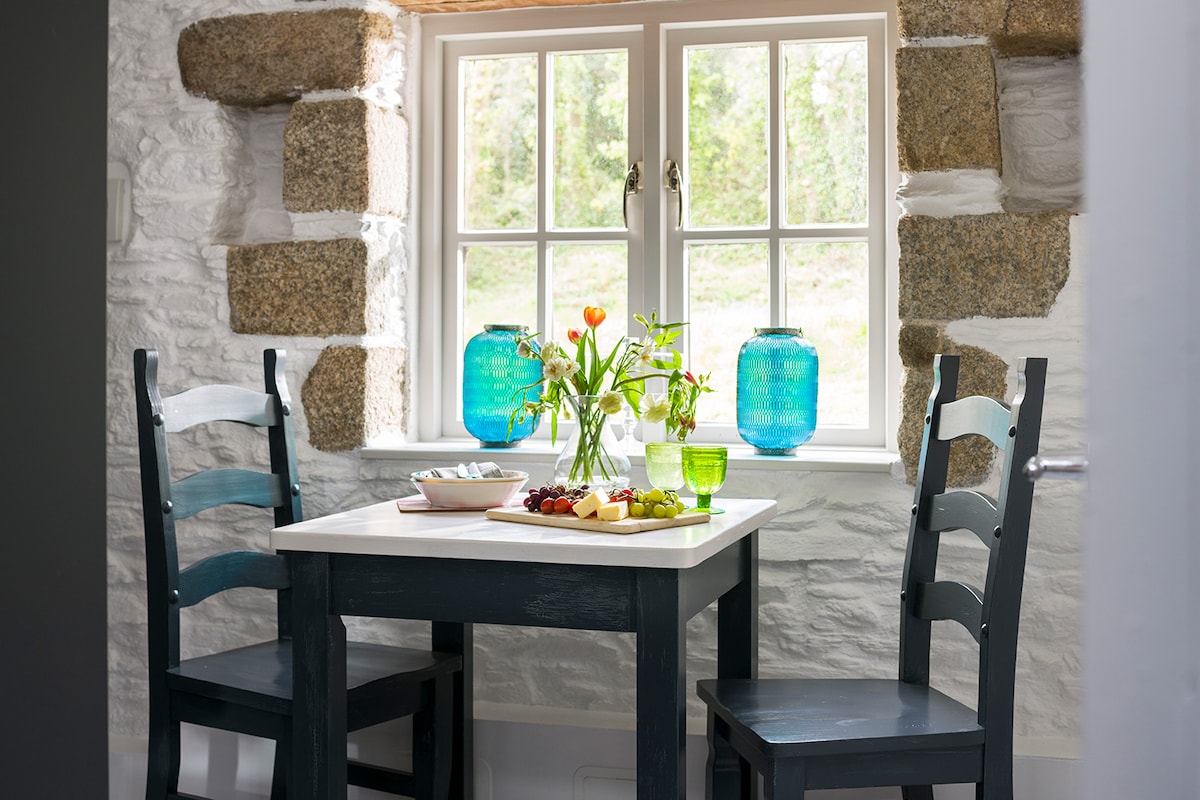 A cozy dining area features a small table adorned with fresh flowers and a selection of fruits. Two dark chairs surround the table, positioned under a window framed by natural stone. Bright blue glass jars add a pop of color, enhancing the sunlit space.