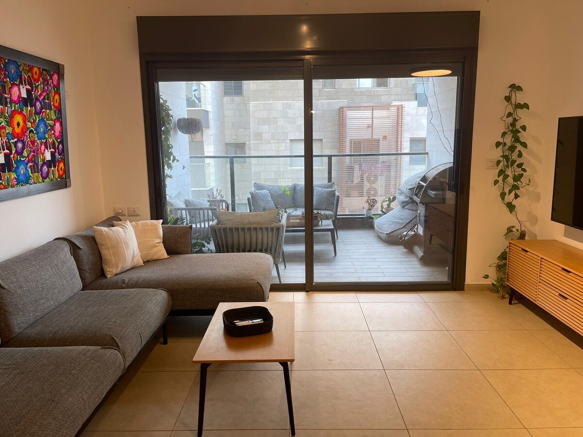 A spacious living area is displayed with a modern couch positioned beside a coffee table. Large glass doors lead to a balcony area, partially revealing outdoor seating. A colorful wall art piece adds a focal point, while greenery climbs along the wall.