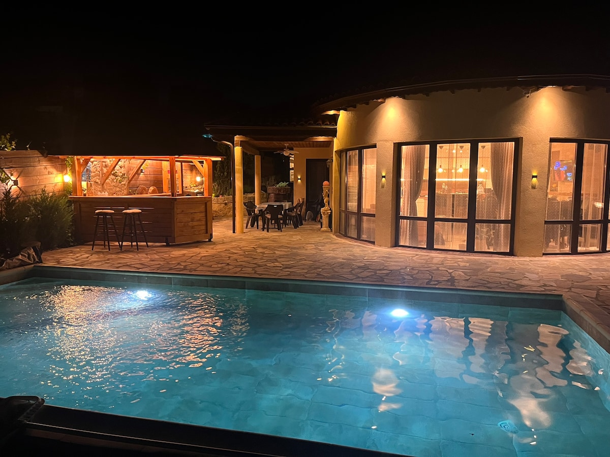 An inviting outdoor area features a pool illuminated by soft lights, alongside a wooden bar with seating. The villa's large windows reflect a warm glow from the interior, while the surrounding space is designed for relaxation and social gatherings.