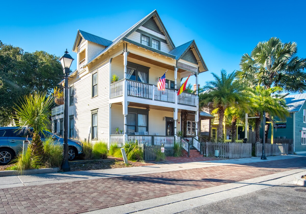 The historic Victorian house is set against a clear blue sky, featuring a classic façade with a prominent balcony. Lush palm trees frame the entrance, while brick paving leads to the front steps. Flags are displayed, emphasizing the welcoming atmosphere of the property.