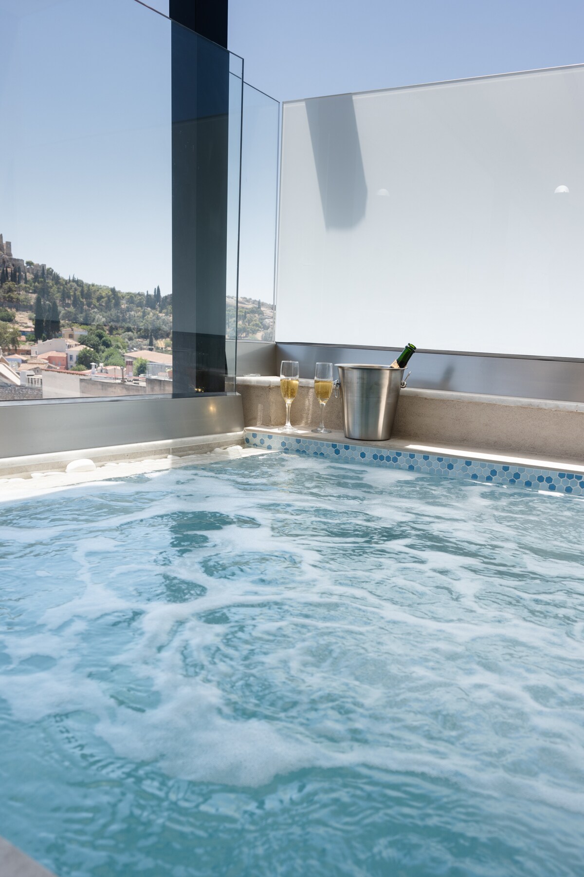 A modern jacuzzi is positioned by large glass windows, allowing natural light to illuminate the space. Bubbles gently rise in the water, complemented by a nearby bucket holding champagne flutes. The backdrop features a view of surrounding hills and architecture.