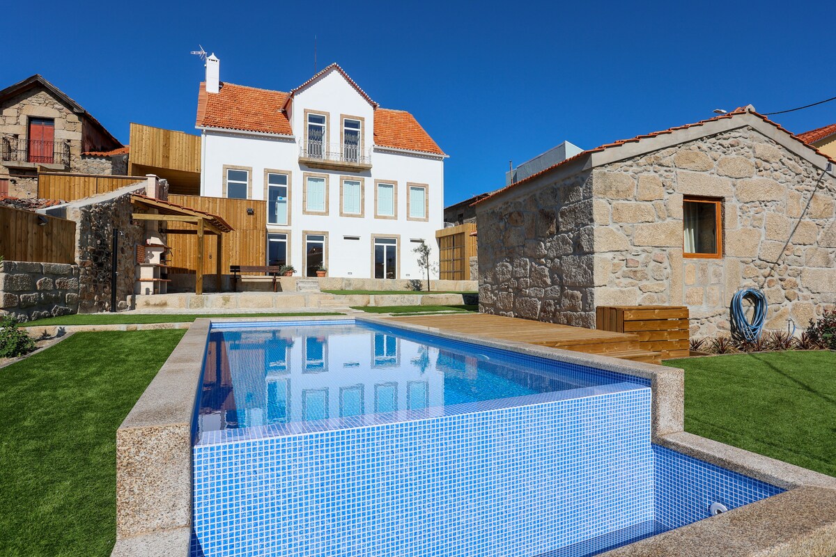 A modern outdoor space features a clear swimming pool surrounded by manicured grass. The main house, with its traditional architecture, includes large windows and a welcoming terrace. A rustic stone structure sits nearby, adding character to the setting.