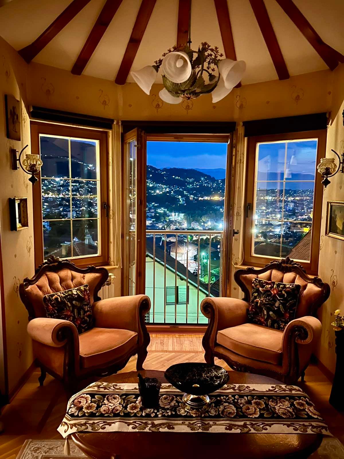 Two vintage armchairs are positioned near large windows, offering a panoramic view of the city lights at night. A small table is placed between the chairs, and decorative elements are visible on the walls, enhancing the cozy atmosphere of the corner space.