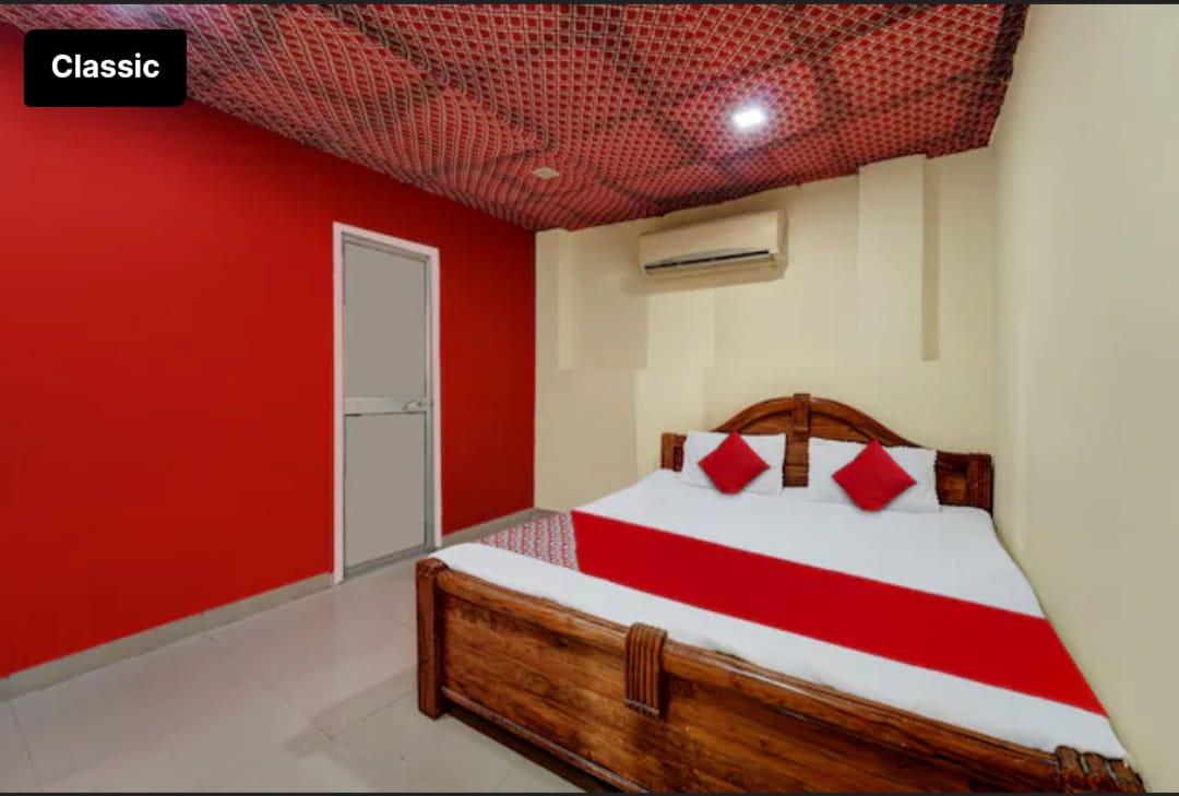 A cozy double room features two double beds, accented with red cushions. The walls are painted in a vibrant red, creating a warm ambiance. An air conditioning unit is installed for comfort, while a light-colored door is visible on one wall, contributing to the room's layout.