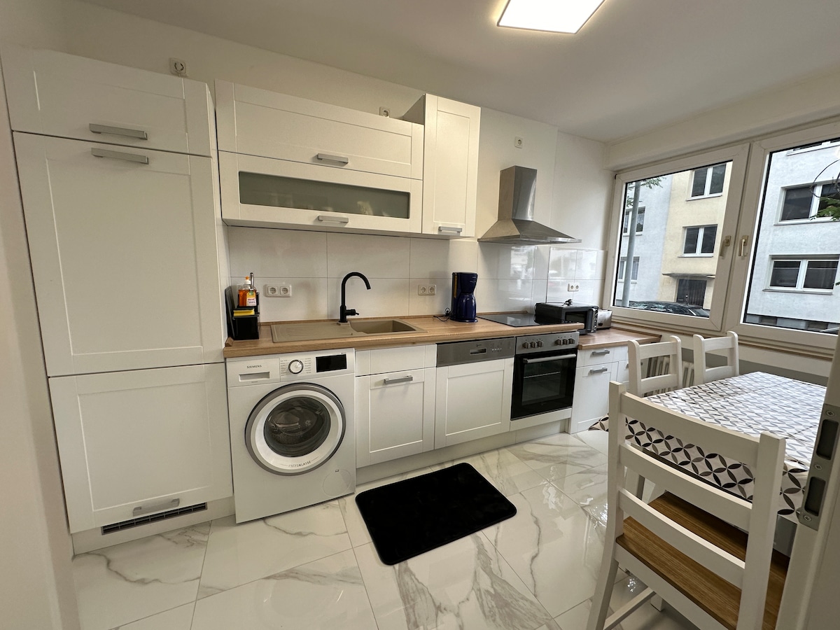 A modern kitchen is showcased, featuring white cabinetry and a wooden countertop. Appliances include a washing machine, stove, and oven. Natural light enters through a large window, illuminating the space. A dining table with white chairs is positioned nearby, enhancing the functional layout.
