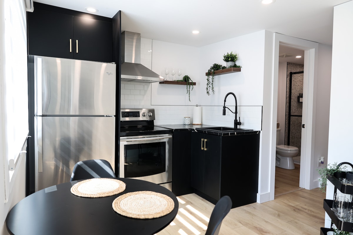 A modern kitchen area features sleek black cabinetry and stainless steel appliances, including a refrigerator and oven. A small round dining table with two place settings is situated near a window, allowing light to brighten the space. Open shelving displays decorative plants.