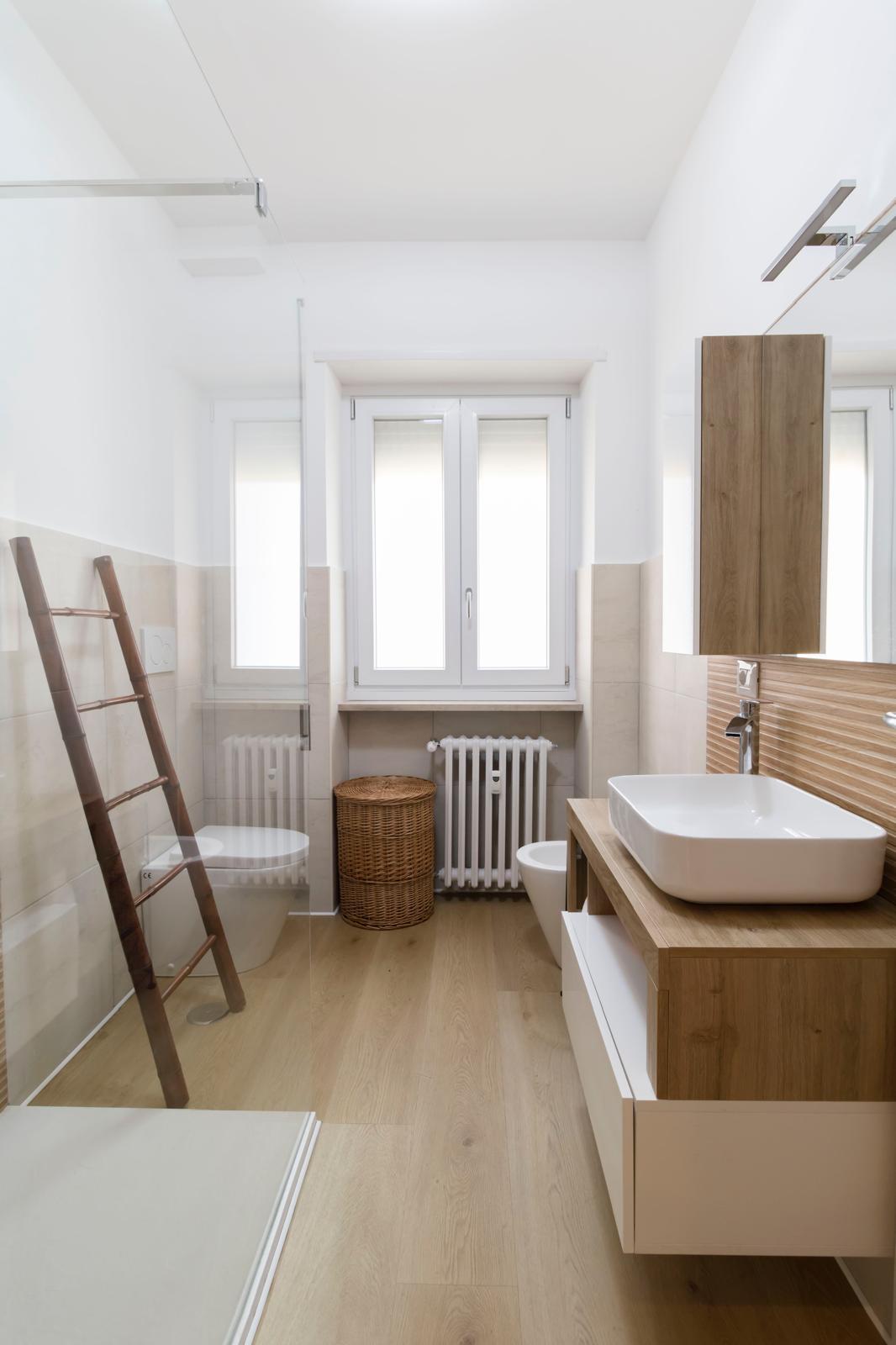 A modern bathroom features a spacious walk-in shower with clear glass. Natural wood accents complement a floating vanity with a white sink. A tall, wooden ladder leans against the wall, and a woven basket is positioned beside the radiator under a window, allowing natural light.