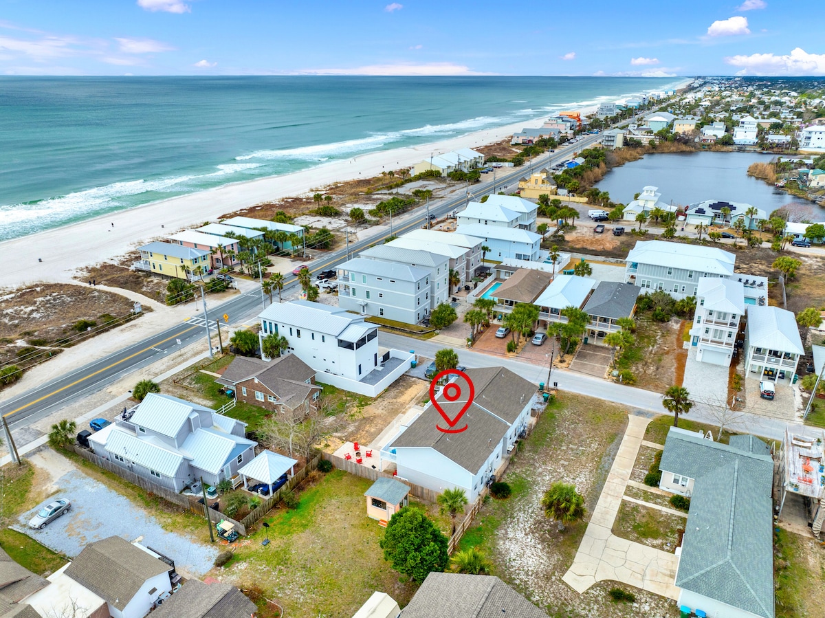 An aerial view captures the surrounding landscape, featuring the coastline and beach area. A mix of residential structures is visible, along with pathways leading to the beach. The location is highlighted with a marker, indicating proximity to the Gulf and local amenities.