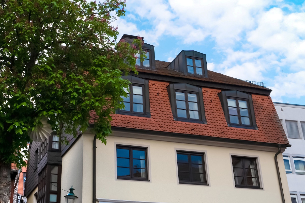 The exterior of a stylish building is shown, featuring a mix of modern and traditional architectural elements. The upper levels are highlighted by dormer windows beneath a sloped, tiled roof. A green tree partially frames the building, complementing the overall aesthetic.
