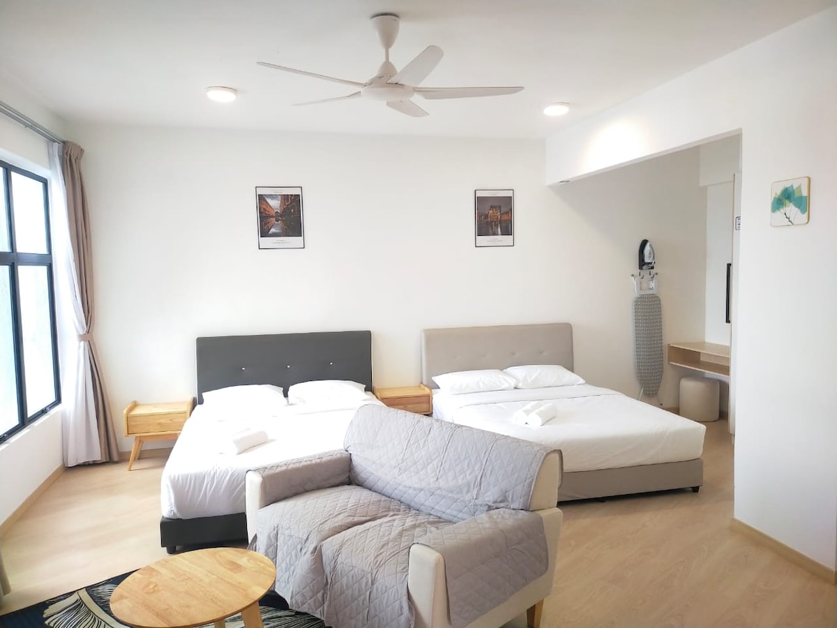 A well-lit bedroom features two spacious beds accompanied by bedside tables. A comfortable sofa sits centrally in the room, with light-colored curtains framing large windows. Two framed artworks are displayed on the wall, complementing the minimalist decor.