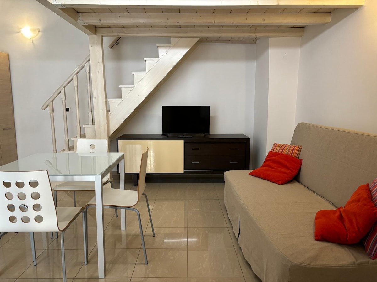 A modern living area features a light-colored sofa with colorful cushions and a dining table surrounded by four chairs. A flat-screen television is positioned on a wooden cabinet, and a staircase leads to the upper level.