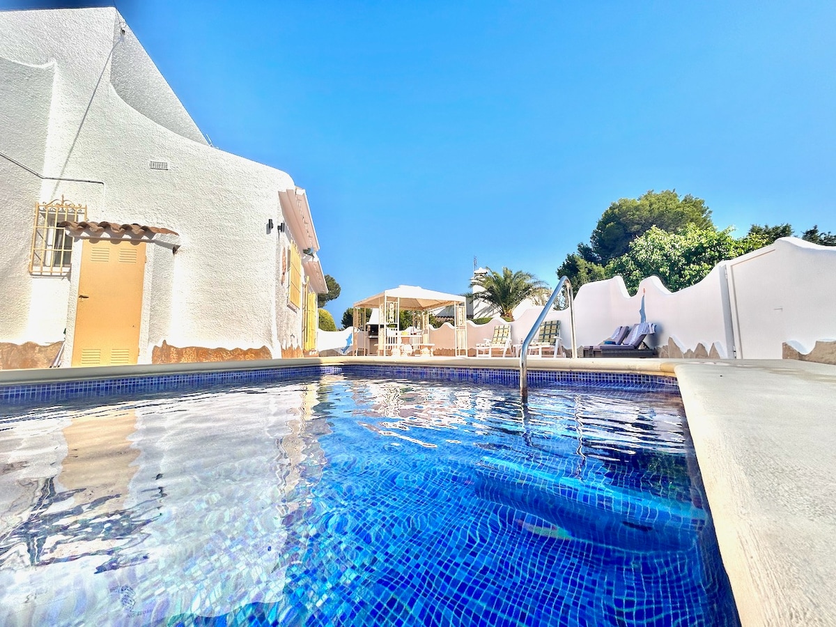 A serene private pool presents crisp blue waters, surrounded by a sun terrace. Comfortable sun loungers and an outdoor dining area under a modern pergola can be seen in the background, framed by green foliage and a clear blue sky.