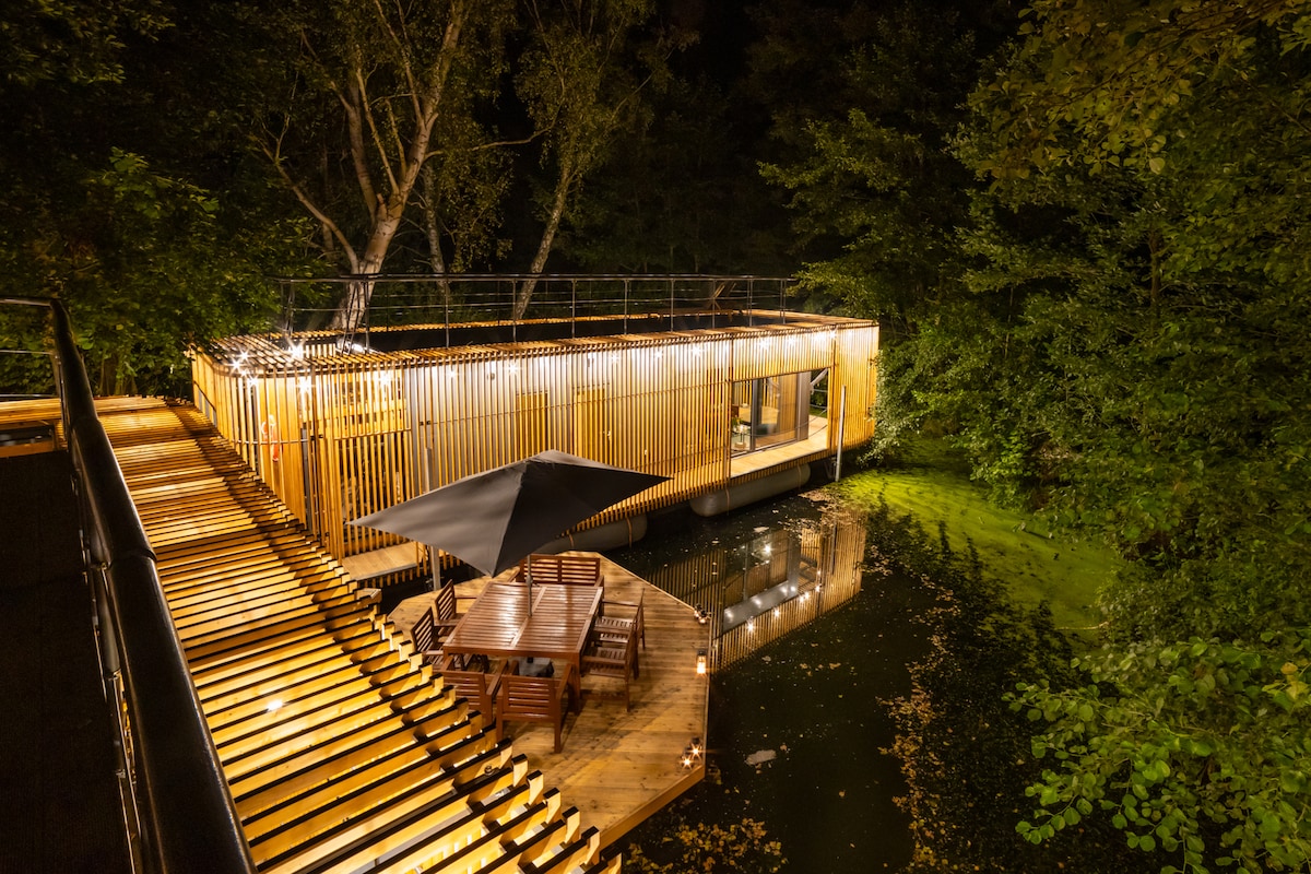 A wooden terrace extends over calm waters, leading to a well-lit floating cabin surrounded by trees. The exterior is adorned with warm lighting, enhancing the secluded ambiance of the space at night. A shaded seating area is visible on the dock, inviting relaxation.