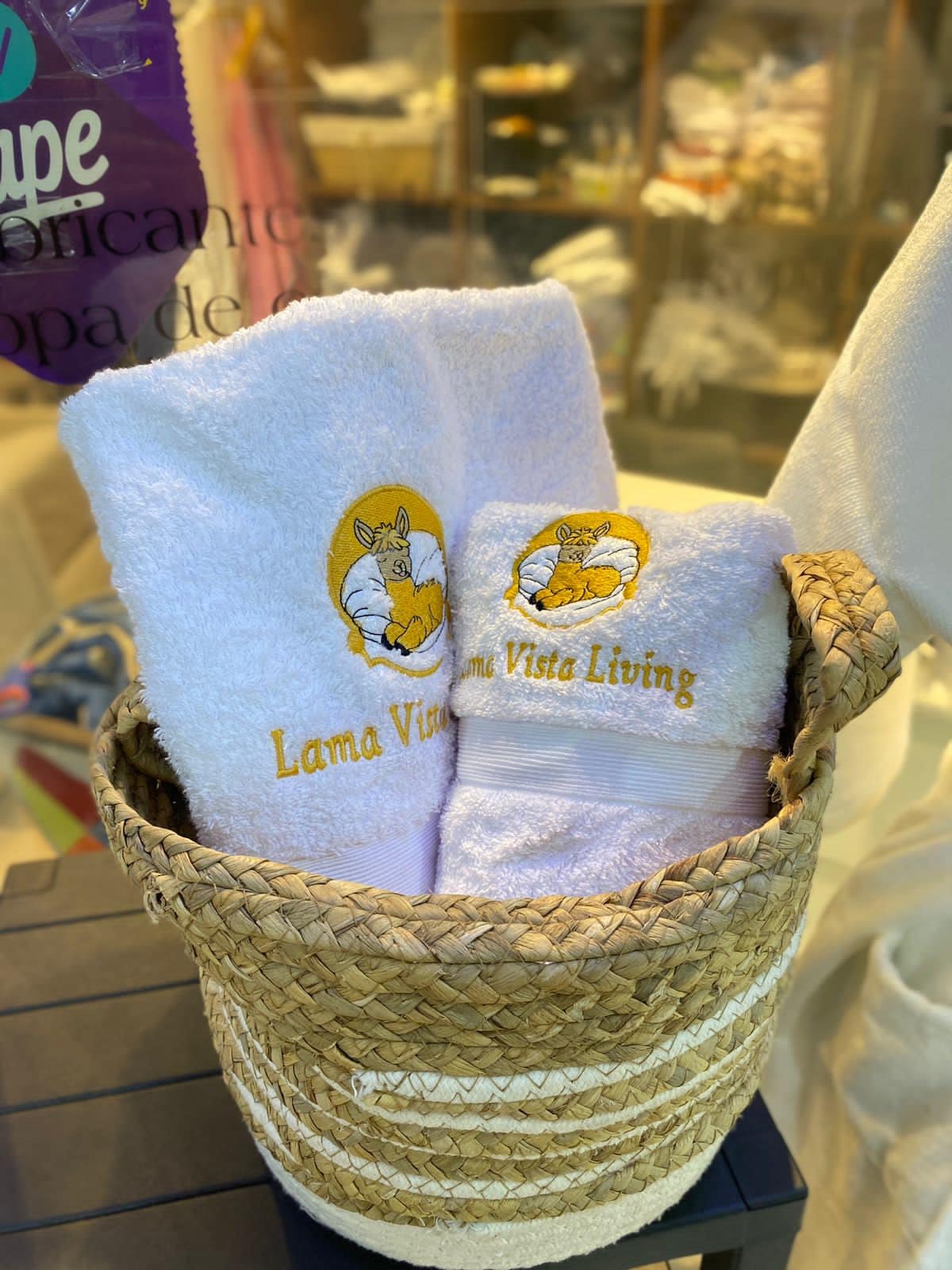 Two neatly folded white towels are displayed in a woven basket. The towels are emblazoned with a logo that reads 'Lama Vista Living,' highlighting the accommodation's identity. The basket provides a rustic touch, complementing the surrounding environment.