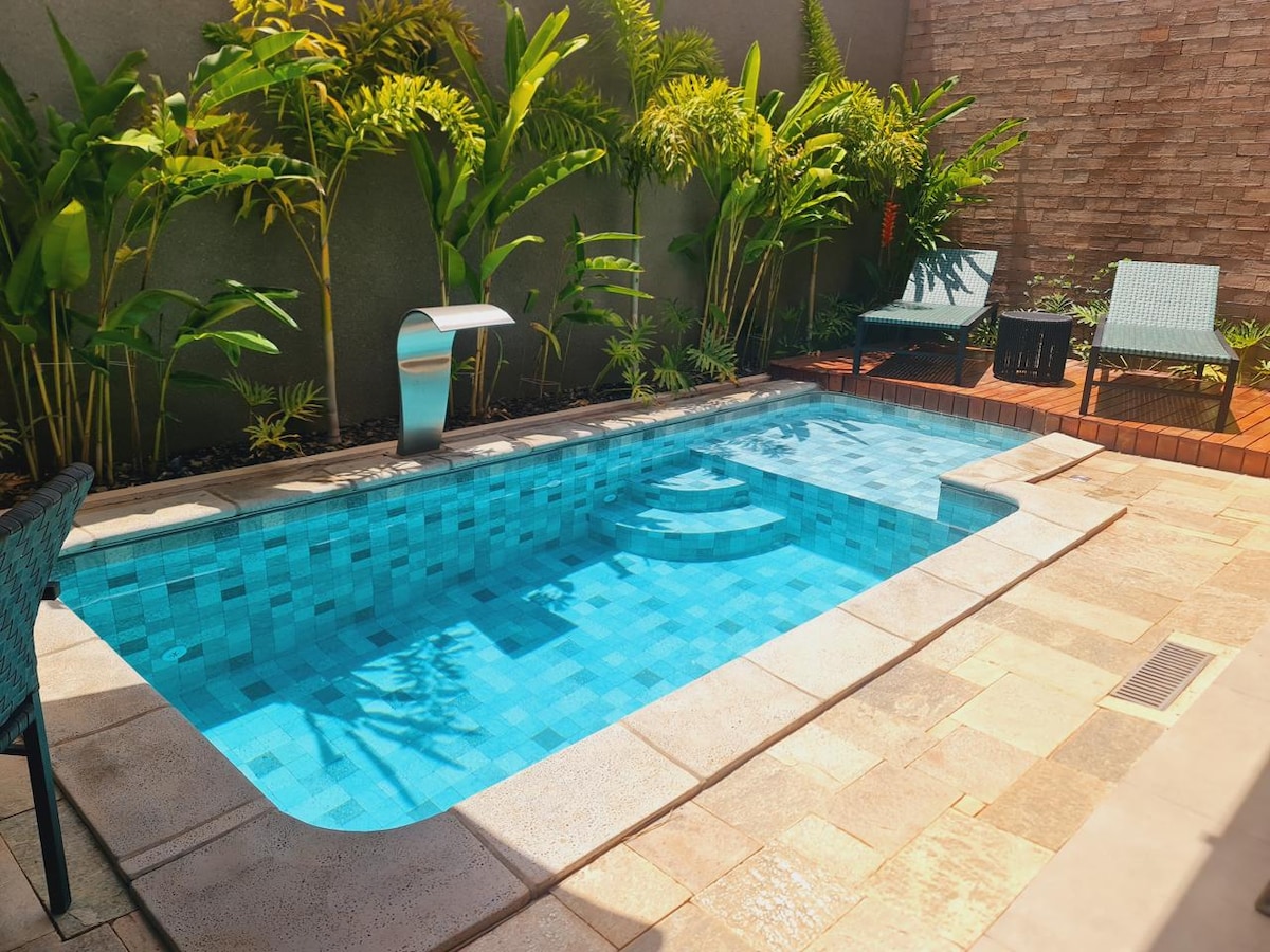 A compact outdoor pool is surrounded by lush green plants, creating a serene environment. Two lounge chairs are positioned nearby, and a small table complements the seating area. The pool’s clear water reflects the sunlight, enhancing the inviting atmosphere.