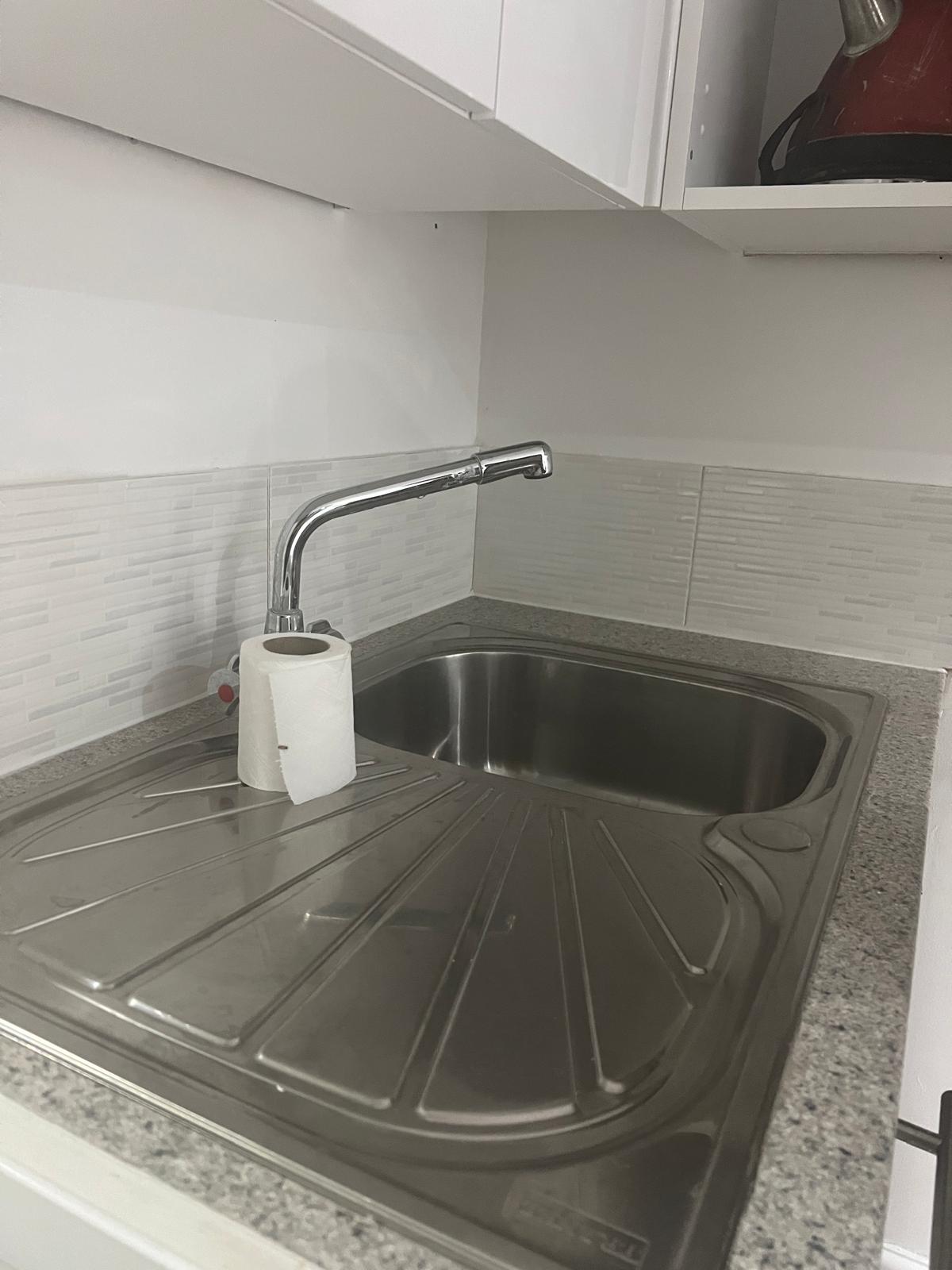 A compact kitchen area features a stainless steel sink set into a gray granite countertop. A roll of paper towels is placed beside the sink. The backsplash consists of light-colored tiles, enhancing the clean and functional design of the space.