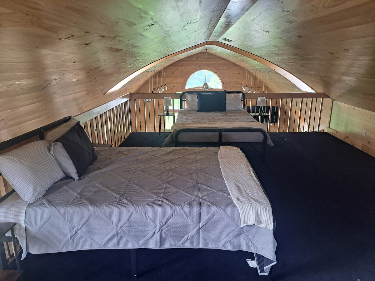 An airy loft area features two beds, each dressed in neutral-toned bedding. Light pours in through an arched window, illuminating the wooden ceiling and walls. A railing surrounds the loft, providing an open view of the space below.