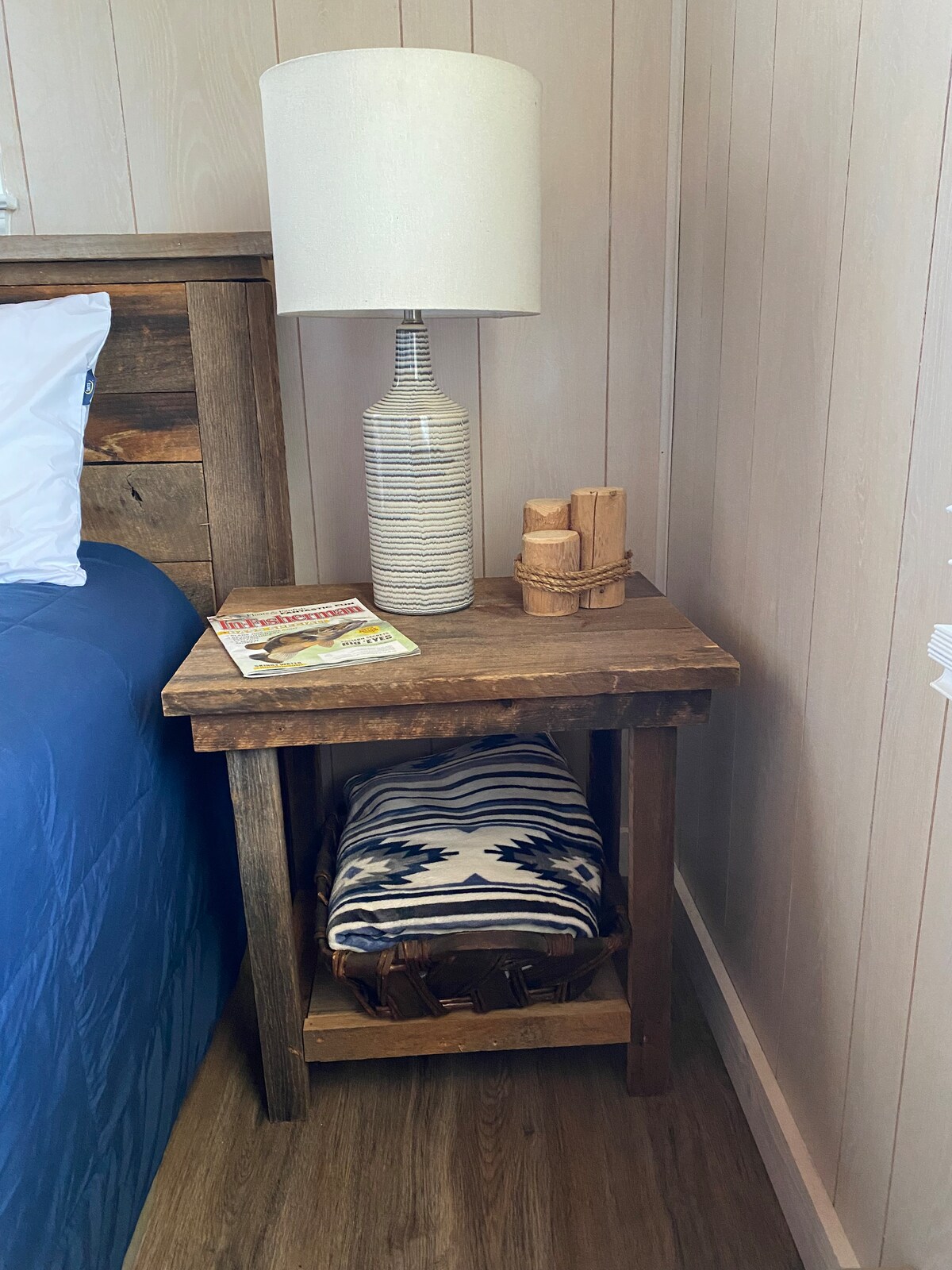 A wooden bedside table is positioned beside a spacious bed, topped with a textured lamp and a stack of magazines. A woven basket is located below the table, covered by a patterned blanket, creating a cozy and organized sleeping area.