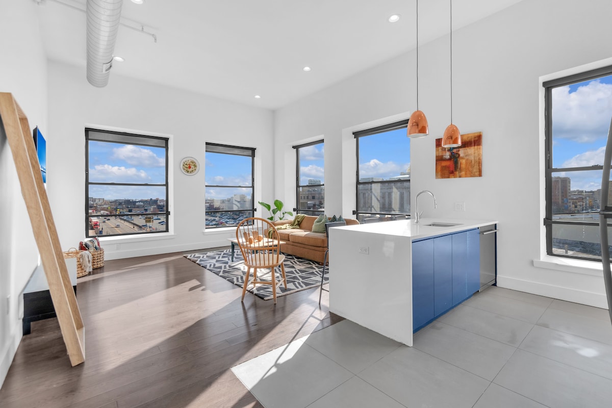 An open design living area features large windows allowing for ample natural light. A modern kitchen with blue cabinetry is complemented by a spacious living area outfitted with a comfortable sofa and decorative rug. Stylish lighting fixtures hang above the kitchen island.