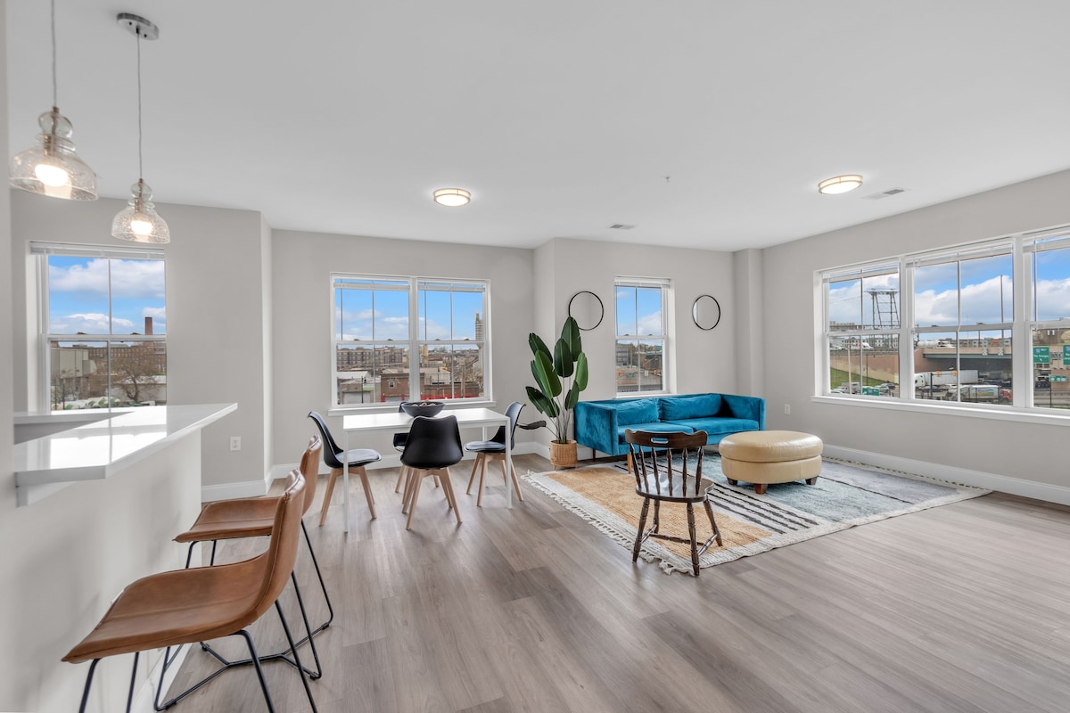 An open-concept living area features large windows that allow natural light to fill the space. A blue sofa contrasts with a light wood floor, while a round ottoman is positioned nearby. A dining table with modern chairs provides a comfortable spot for meals, and two hanging lights illuminate the area.
