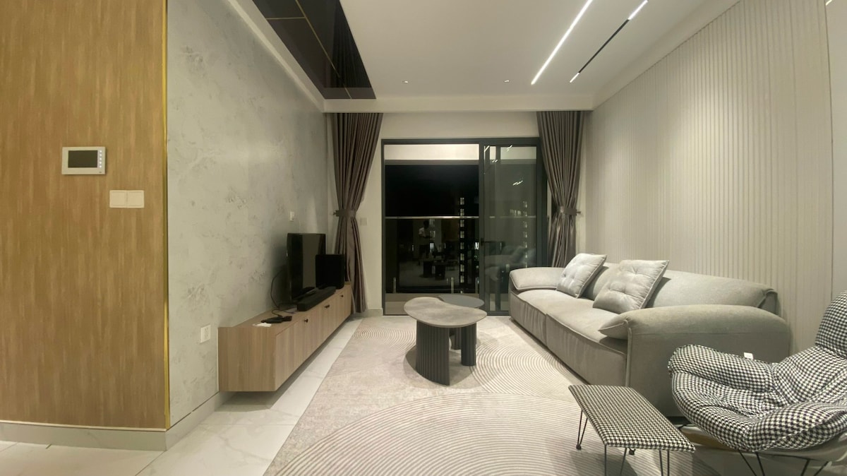 A modern living area features a comfortable gray sofa and a round coffee table. A media console is positioned below a wall-mounted TV. Natural light enters through large glass doors, which are framed by floor-length curtains, highlighting the soft color palette and contemporary design.