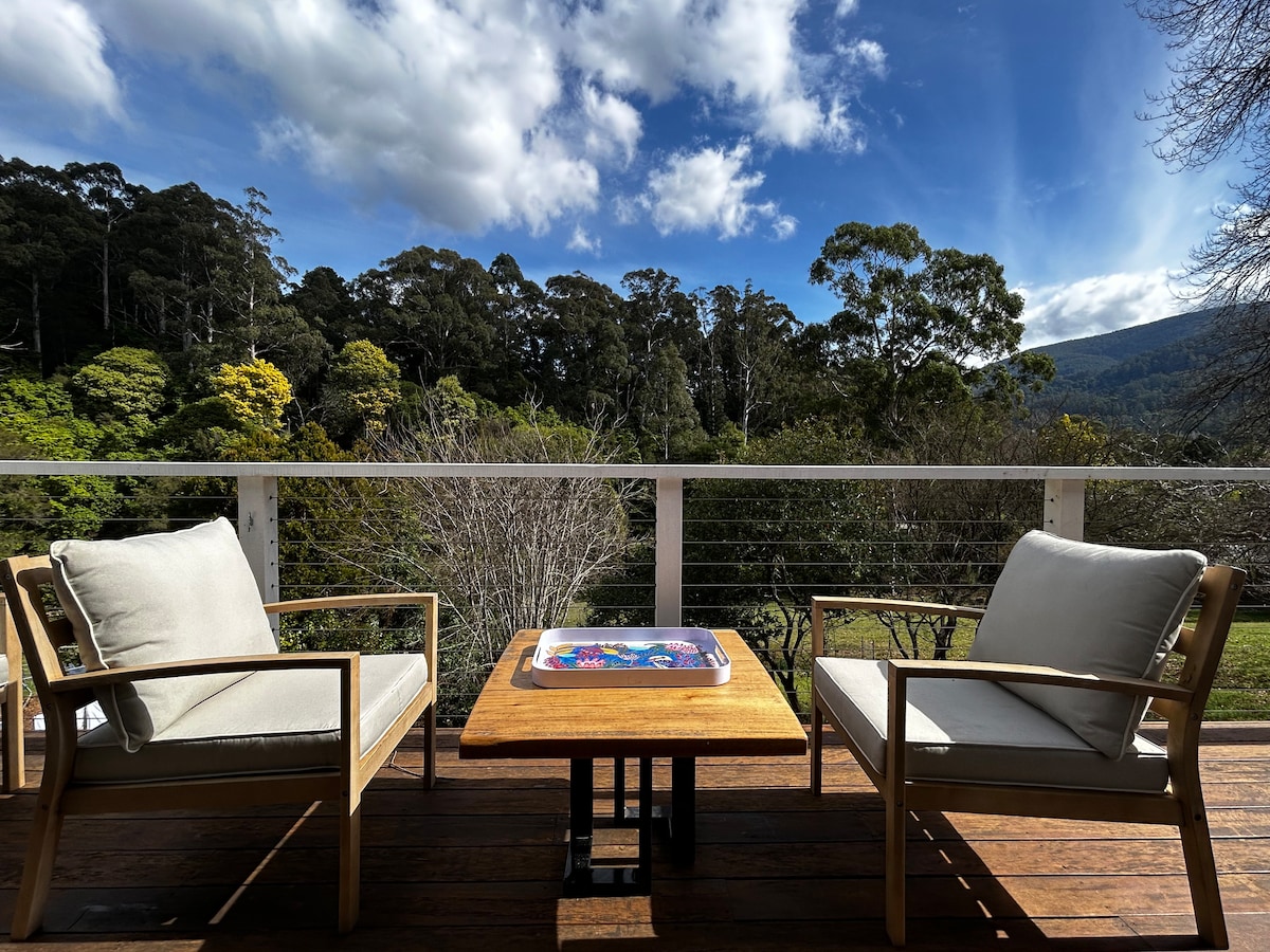 A spacious balcony features two comfortable seating chairs placed around a wooden table. Lush greenery and trees provide a scenic backdrop under a partly cloudy sky, creating a serene outdoor space ideal for relaxation.