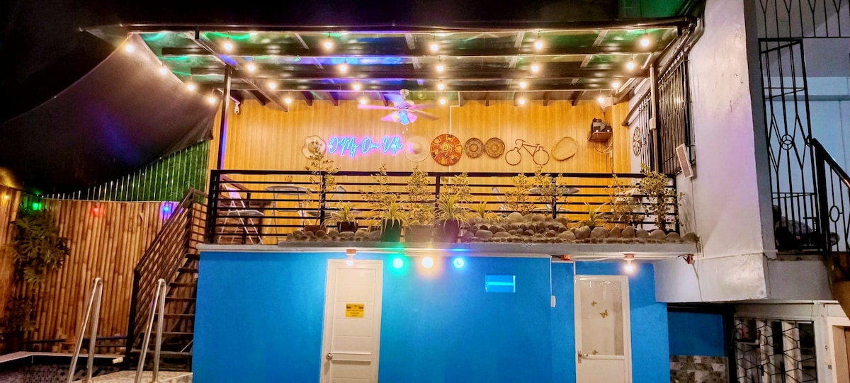 An inviting exterior of a villa is illuminated with decorative lights, highlighting the blue walls and wooden accents. A spacious deck features potted plants and wall decorations, creating a welcoming entrance. Stairs lead to the upper level, enhancing the villa's appeal.