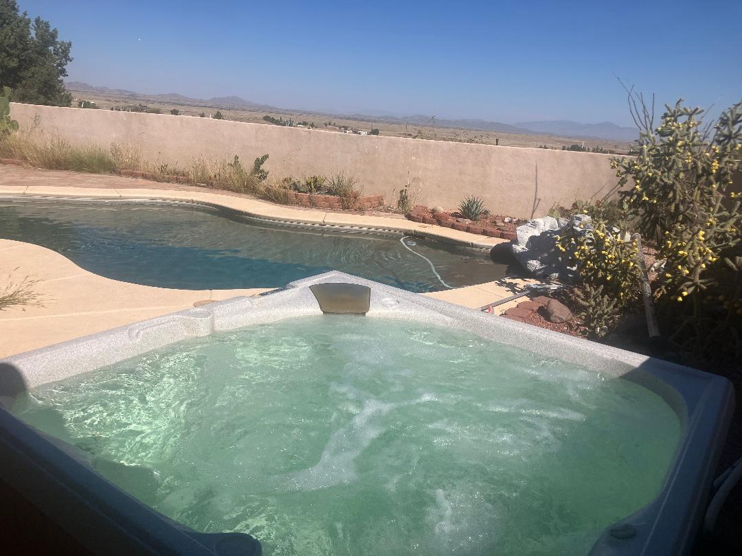 A hot tub is featured in the foreground, offering a view of a swimming pool and surrounding desert landscape. The area is bordered by a low wall, and various desert plants can be seen in the background under a clear blue sky.
