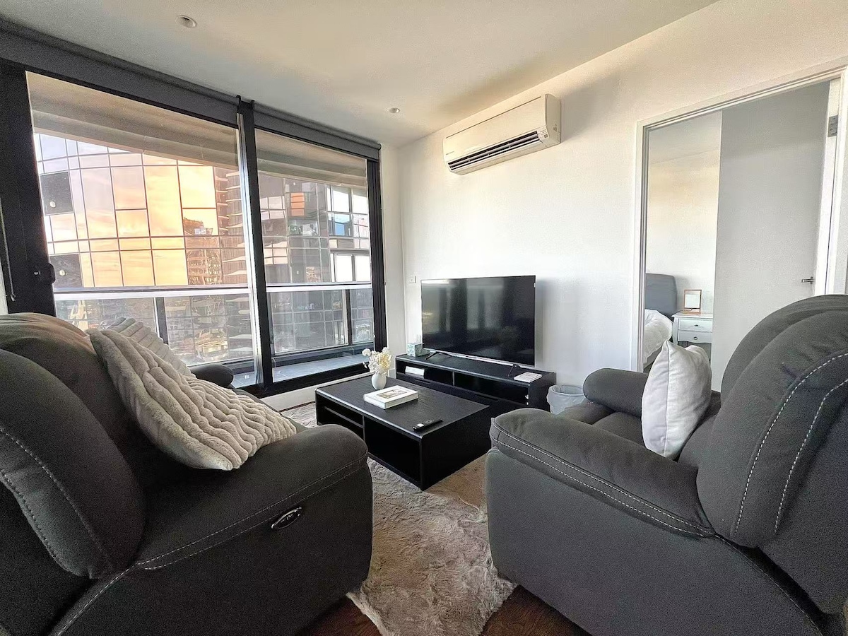 A cozy living area features dark sofas with soft cushions, positioned around a sleek coffee table. A large window allows natural light to fill the space, while a smart TV sits on a low entertainment unit. A mirror reflects the room's contemporary design.