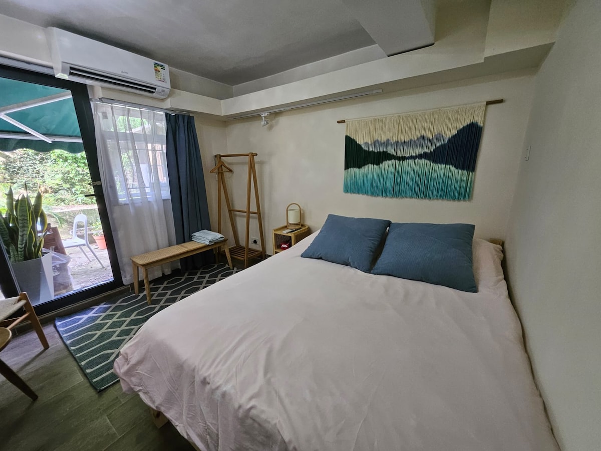 A spacious sleeping area features a large bed with two blue pillows. A hanging wall tapestry with mountain designs adds a touch of color. Natural light enters through sliding glass doors, which lead to a serene outdoor space filled with greenery.