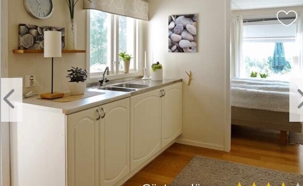 A well-lit kitchen area features white cabinetry and a dual sink. Natural light flows through the windows, illuminating a serene bedroom in the background. A decorative piece of smooth stones is displayed on the wall, complemented by soft textiles and a warm wood floor.