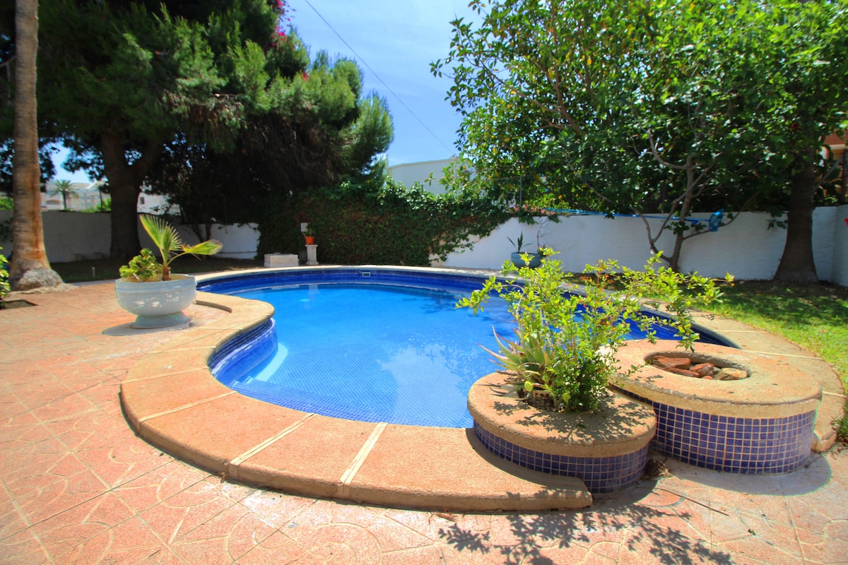 A private swimming pool is surrounded by lush greenery and features a stone coping. Several potted plants are placed along the edge, adding natural elements to the tranquil outdoor space. Sunlight reflects off the clear water, creating a relaxing ambiance.