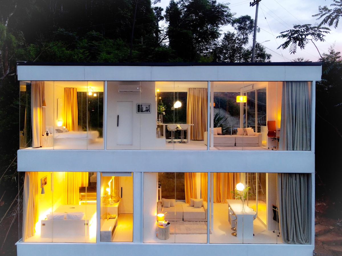 A modern glass structure is highlighted by expansive windows showcasing a well-lit interior across three floors. Warm light emanates from multiple rooms, creating a welcoming ambiance. Surrounding greenery complements the home, providing a connection to nature.