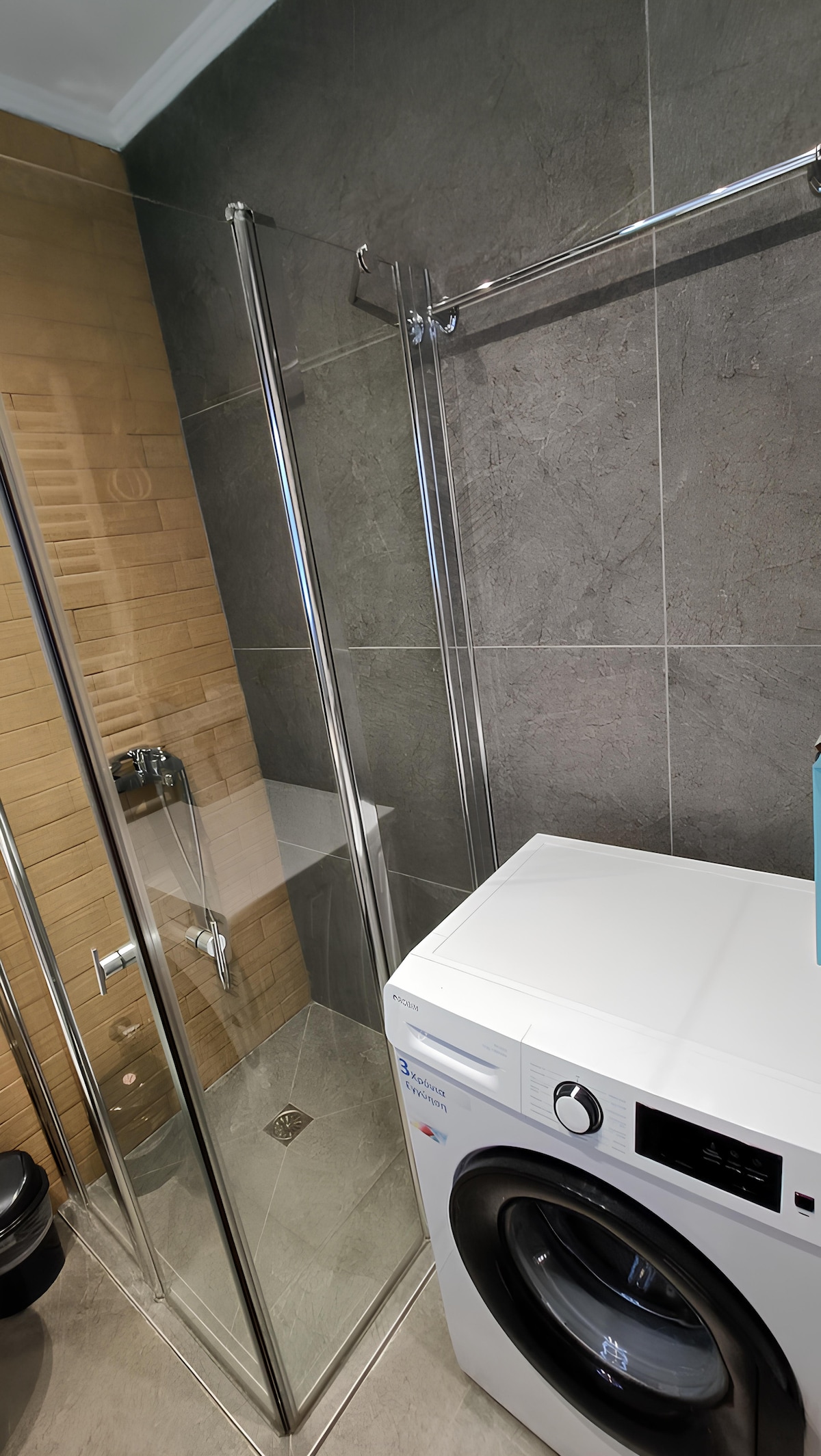 The bathroom features a glass shower enclosure alongside a modern washing machine. The walls are finished with a combination of smooth gray tiles and textured brown accents. A compact space is effectively utilized, creating a functional area with necessary amenities.
