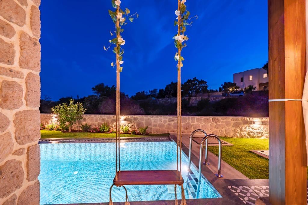 A private pool glistens under the evening sky, framed by lush greenery. A swing hangs nearby, adding a playful touch. Soft outdoor lighting enhances the tranquil atmosphere, while modern stone architecture complements the natural surroundings.